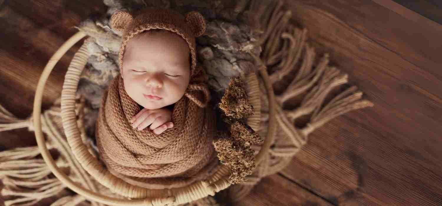 Senso-Blanky Hero Baby wrapped in brown knitted swaddle and wearing a bear hat in a wooden basket.