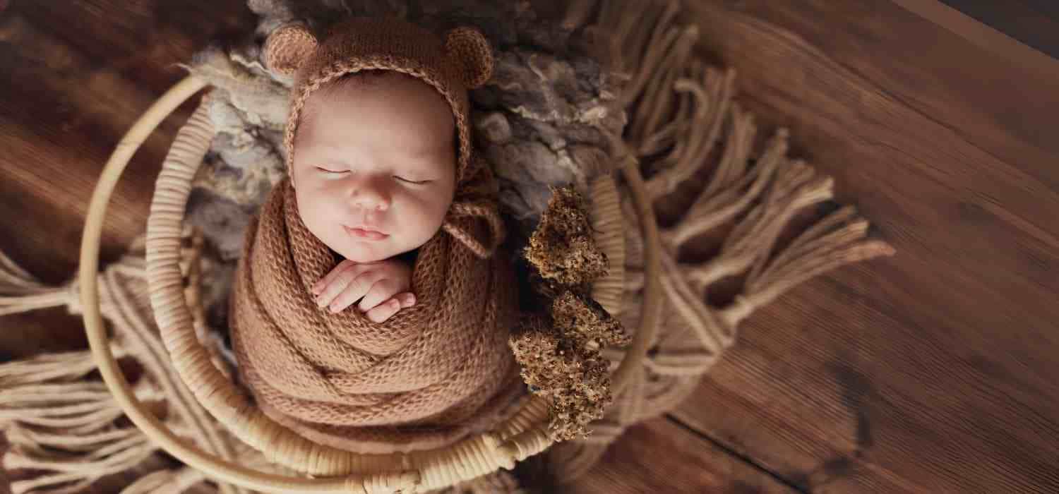 Senso-Blanky Baby wrapped in brown knitted swaddle and wearing a bear hat in a wooden basket.