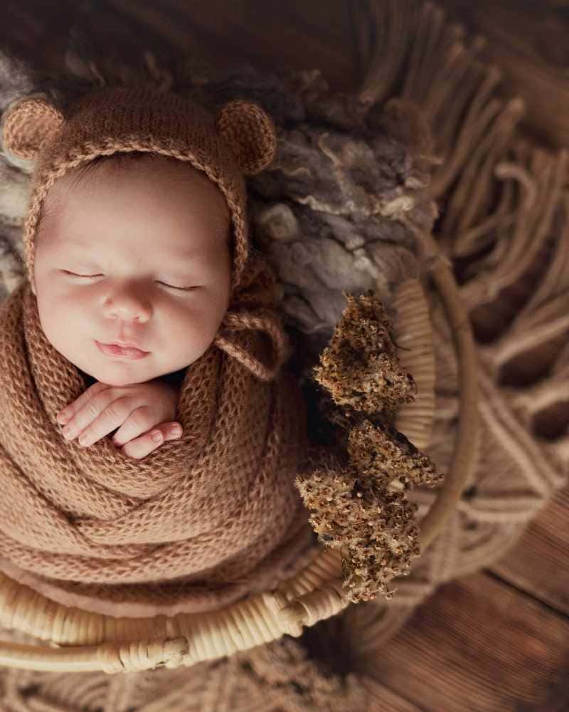 Senso-Blanky baby wrapped in brown knitted swaddle blanket and wearing a matching brown bear hat in a textured basket.