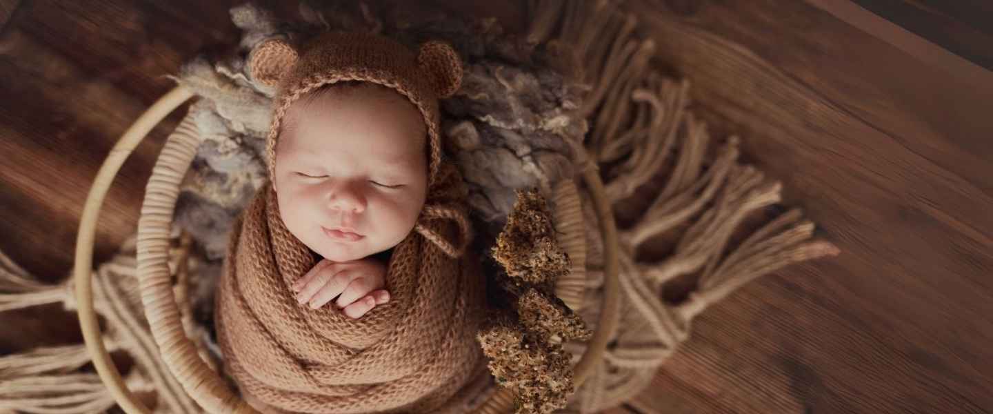 Senso-Blanky Hero Baby wrapped in brown knitted swaddle and wearing a bear hat in a wooden basket.