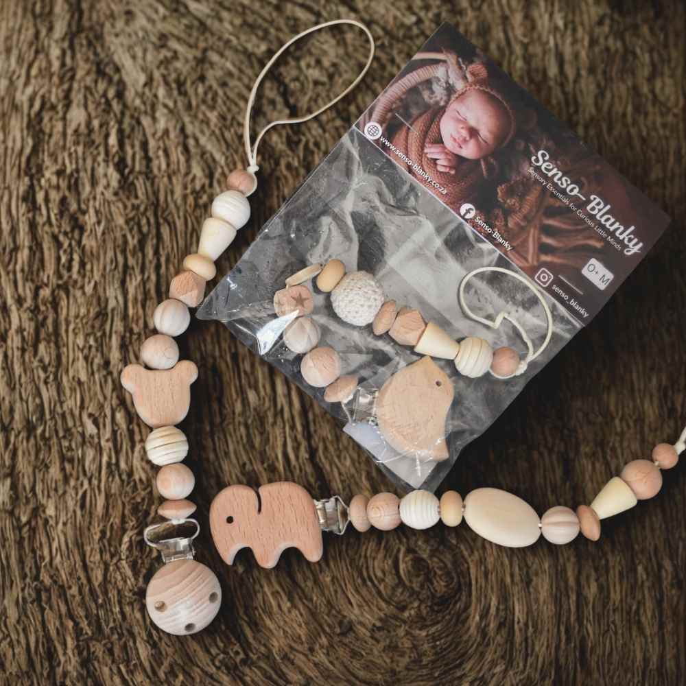 Senso-Grip premium baby dummy clip beautifully displayed in its retail packaging, resting on natural tree bark background for a rustic, organic feel. Handcrafted sensory pacifier chain by Senso-Blanky South Africa.