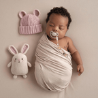 A sleeping newborn baby swaddled in a premium muslin cloth, wearing a soft pink beanie cap. The baby has a pacifier in mouth, secured by a handcrafted dusty rose Senso-Grip dummy clip attached safely to the swaddle. A plush bunny toy rests gently in the soft-focus background of this cozy, teacher-approved sensory setting.
