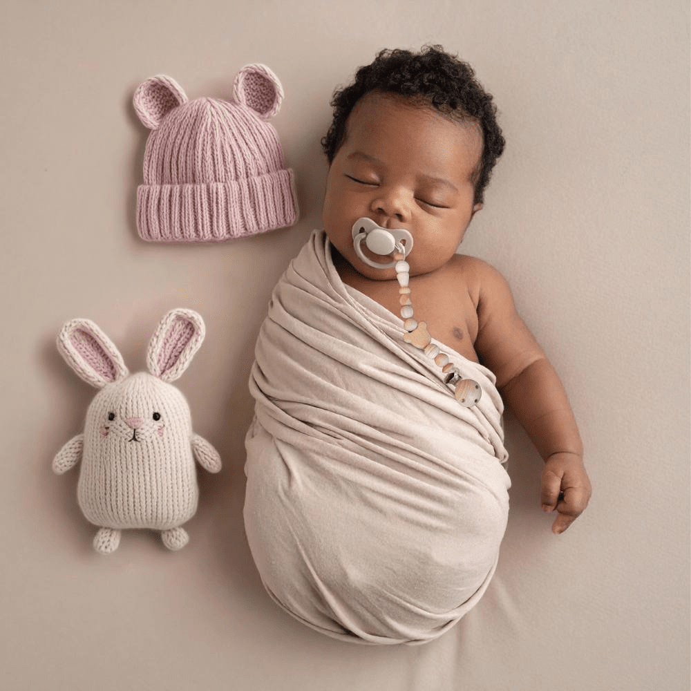 A sleeping newborn baby swaddled in a premium muslin cloth, wearing a soft pink beanie cap. The baby has a pacifier in mouth, secured by a handcrafted dusty rose Senso-Grip dummy clip attached safely to the swaddle. A plush bunny toy rests gently in the soft-focus background of this cozy, teacher-approved sensory setting.